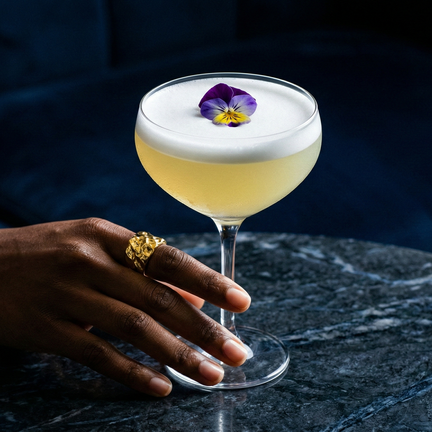 Cocktail with a decorative flower on a dark surface, with a hand resting on the table.